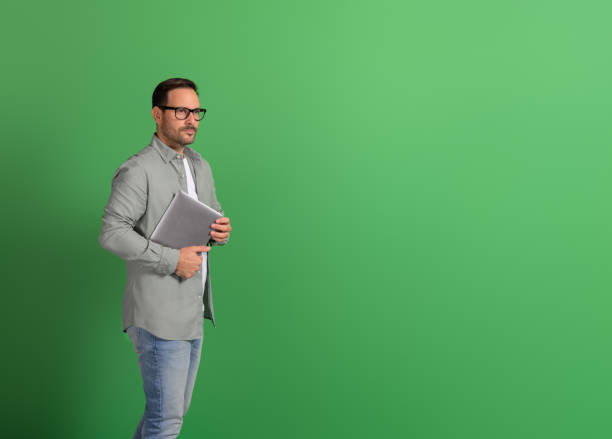 istock Pensive young businessman holding wireless computer and looking away seriously on green background 2151088159