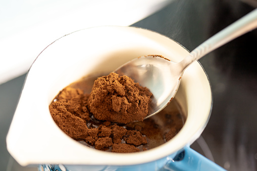 Gently, a spoonful of aromatic ground coffee is deposited into the swirling, boiling water. As it dissolves, the kitchen fills with the rich scent, promising a flavorful brew.