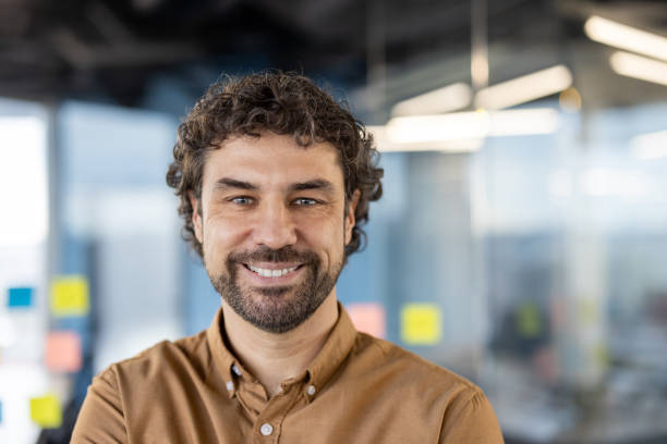 istock Attractive unshaved man with curly hair looking confidently at camera with wide smile on blurred background. Satisfied entrepreneur spending busy day in company building and enjoying working process 2151088883