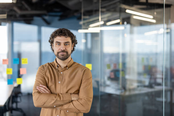 istock Emotionless mature man with arms folded on waist looking at camera while standing in boarding room. Strict boss waiting for employees for discussing important business project and profit increasing 2151090082