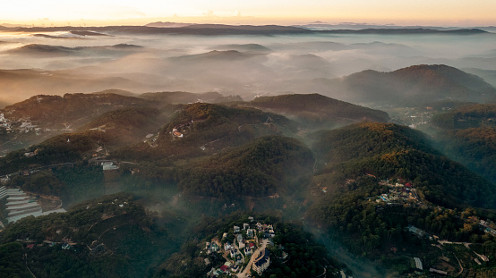 Aerial view of Dalat City at dawn, showcasing mist-covered mountains, lush greenery, scattered buildings, and serene atmosphere creating a picturesque landscape