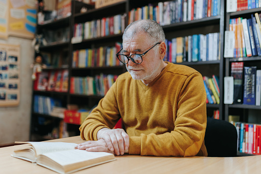 Amidst the tranquility of the bookstore, a white Caucasian American senior man, with glasses and a beard, loses himself in the pages of a book, demonstrating the enduring allure of reading.