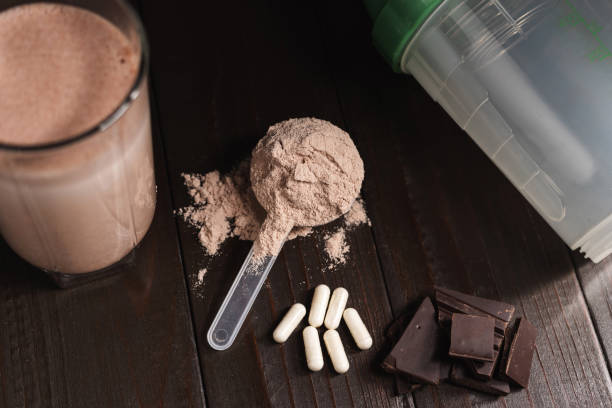 istock Plastic measuring spoon with whey protein powder, milkshake cocktail in a glass, blended protein drink, white pills or capsules, chocolate cubes on a dark wooden background 2151112193