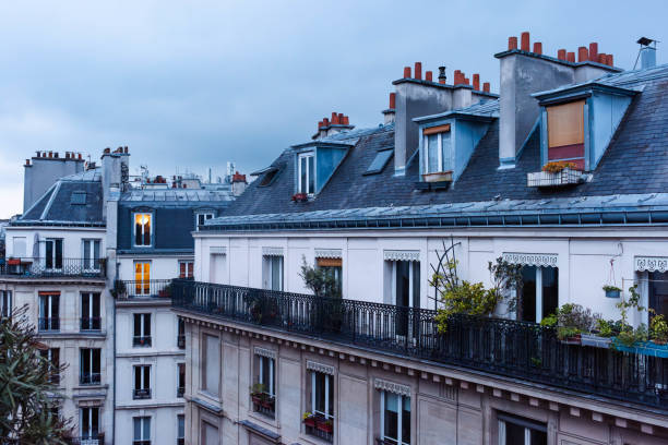 Captured from a fifth-floor balcony, this scene offers a mesmerizing view of Parisian rooftops as evening descends. Chimneys punctuate the skyline, while illuminated windows cast an orange glow against the backdrop of a threatening sky. Experience the atmospheric charm of Paris before nightfall in this urban landscape