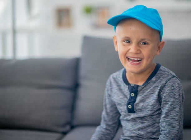 A little boy battling cancer sits at home in between treatments.  He is dressed comfortably and is wearing a blue hat to keep his shaved head warm.