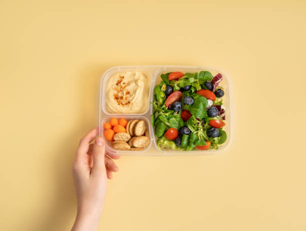 istock Hand holding a  plastic container with hummus and mixed salad on yellow background 2151142083