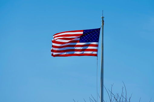 A United States flag attached to a flagpole waves in a strong wind on a sunny day.