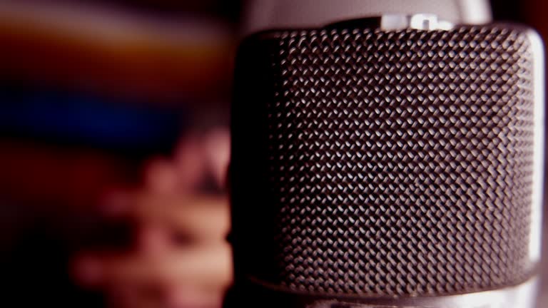Podcast day. A close-up shot of a microphone in a studio. Slowly zooming out on a person recording a podcast in a colorful studio. Recording audio in a soundproof room. A recorder. Person speaking into a microphone.