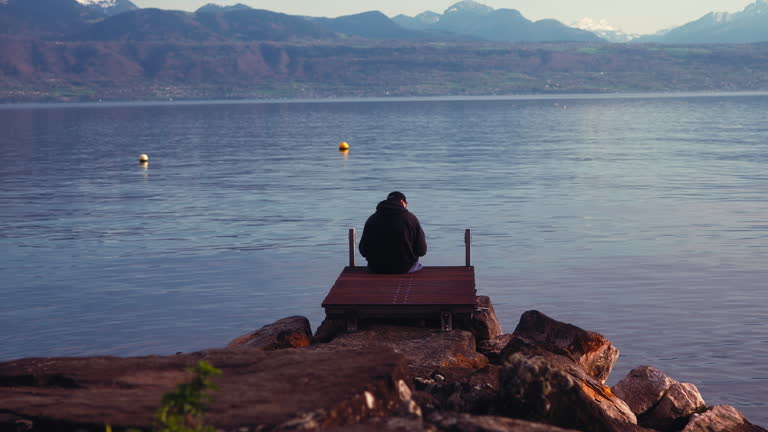 Wide shot of man sitting on a wooden platform at the lake Geneve (lac Lémanz) in Lausanne, Switzerland