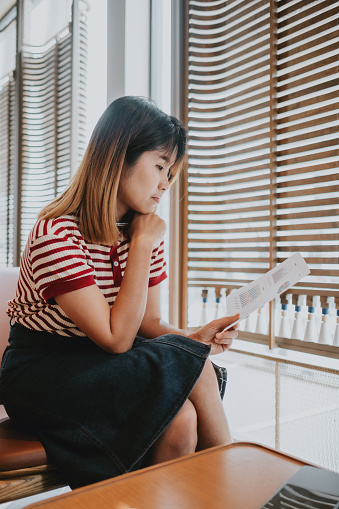 Young Asian woman in a striped shirt reading a financial paper attentively in a well-lit home office.