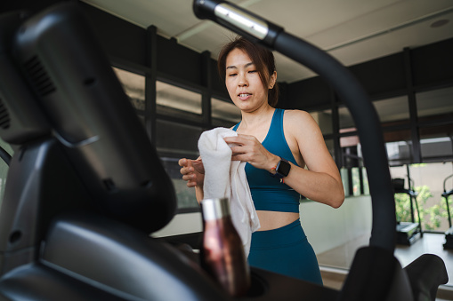 An Asian woman takes a break from running on the treadmill in a fitness gym, wiping away sweat with a towel after her workout.
