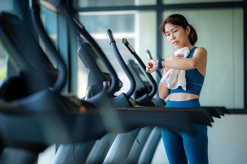 An Asian woman looks at her smartwatch while taking a break from running on the treadmill in a fitness gym, wiping away sweat with a towel after her workout.