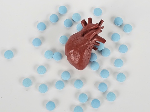 A heart shape is surrounded by numerous blue pills on a white surface. The pills create a contrasting background to the red heart, symbolizing health and medication.