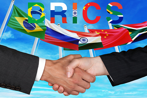 Two hands shaking in agreement with the BRICS countries' flags in the background, symbolizing international cooperation and unity.