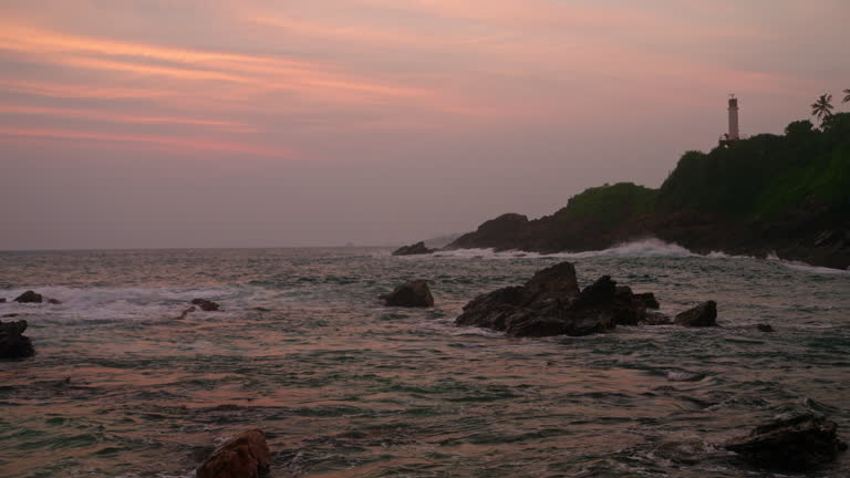 Sunset paints sky over rocky coast, waves crash against stones. Lighthouse stands on cliff, serene evening at sea landscape. Nature scene, tranquil ocean at dusk, coast light beacon guides boats.