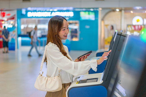 Travel Ready: Young Asian Woman Holds Phone, Checks Travel Info with Passport In Front the Airport Flights Information Display