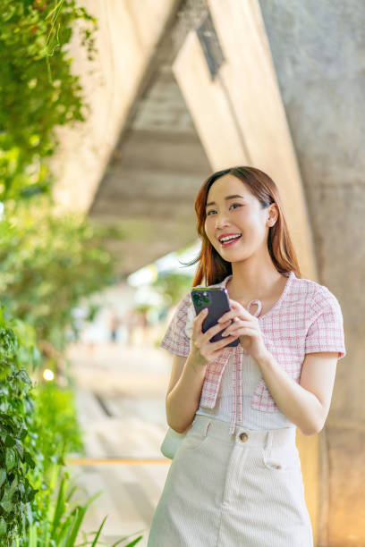 istock Sunlit Connection: Young Asian Woman Uses Phone on Footpath 2151228665