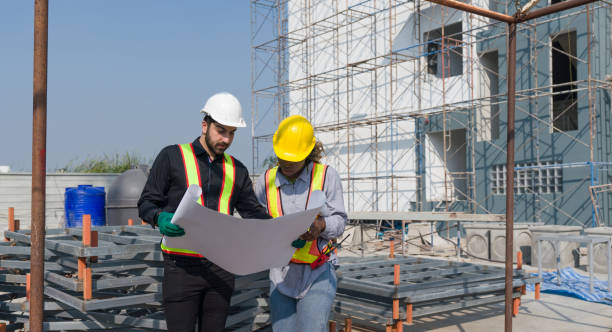 istock Two construction worker looking at a floor plan, standing in front of a large building site with helmet and safety gear. Day time work safety check. Work environment at the site of housing projects. 2151234943