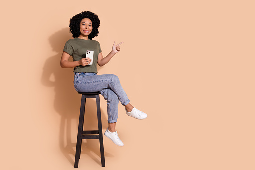 Full size photo of smart person wear t-shirt on chair hold smartphone indicating at logo empty space isolated on pastel color background.