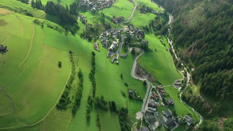 Overhead aerial tilt-down view captures cars navigating a winding road in the quaint village of La Val, South Tyrol, Italy. Lush green hills flank the picturesque scene. LuPa Creative.