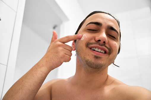 Cheerful white young male with long hair looking at mirror with smile and spreading moisturizing cream on cheeks during skin care routine against white background