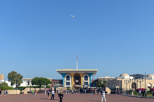 Muscat, Oman - January 2, 2024: Visitors explore the majestic Al Alam Palace square under the clear blue sky as a plane ascends in the background.
