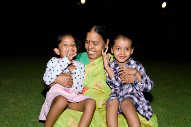 A content Indian grandmother smiles as she embraces her two granddaughters on a cozy blanket in their backyard garden at night