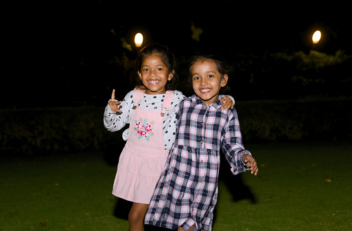 This image depicts two cheerful sisters enjoying a playful evening at a park illuminated by night lights.