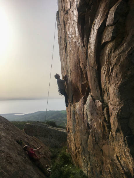 Mature man climbs cliff face above sea and fields, belayer holds rope