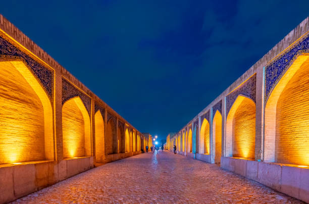 Picture of the iconic bridge of Si o Seh, in the center of Isfahan with iranians standing and relaxing on it. The river Zayandeh is dry due to the season. The Allahverdi Khan Bridge, it is the largest of the eleven historical bridges on the Zayanderud river, It is a popular recreational gathering place, and is one of the most famous examples of Iran Safavid architecture.