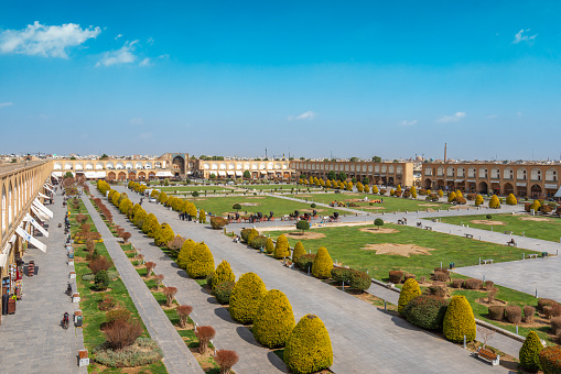 It's a peaceful day at Imam Square (Naqsh-e Jahan Square) as visitors take a leisurely stroll amidst the lush greenery. The historic buildings in the background add to the charm. Isfahan, Iran.