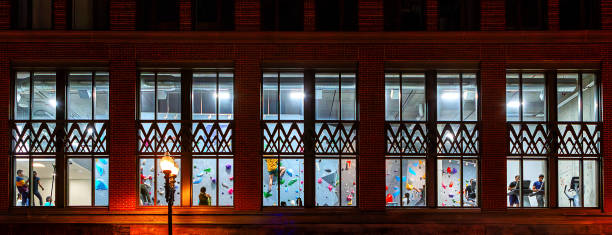 Cambridge, Massachusetts, USA - May 1, 2024: View through the windows of a rock climbing and exercise gym building at night. Young adults climbing and exercising.