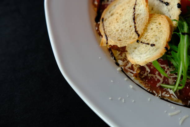 Beef Carpaccio cold appetizer with parmesan, tomatos and arugula on white plate with hardtack on dark background.