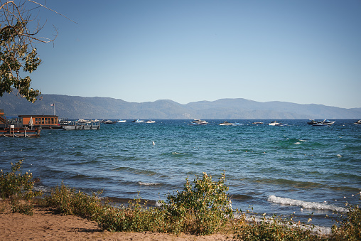 Serene Tahoe lakeside scene with sandy beach, people lounging and walking, blue lake with boats, mountain range backdrop, lush greenery. Ideal for relaxation and recreation.