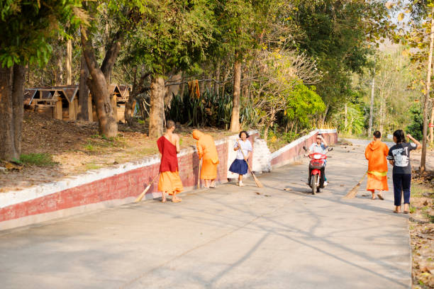 View along street with cleaning young thai monks near and outside of temple Wat Phra Phutthabat Huai Tom in Li District, Lamphun. Street is leading uphill. A young woman and a school girl in school uniform are helping cleaning street with brooms. A person is driving motorcycle. In background are many trees