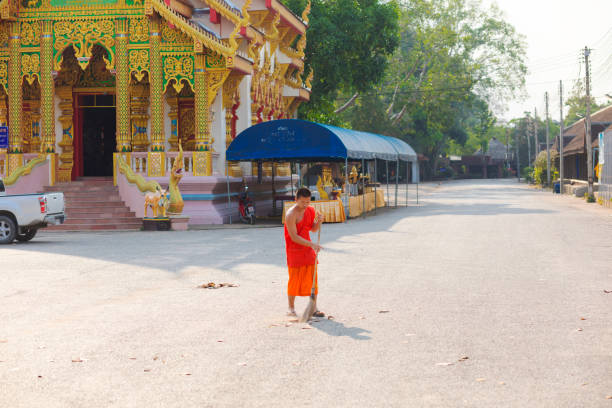 Thai monk is cleaning grounds of temple with broom. Scene is at temple Wat Phra Phutthabat Huai Tom in Li District, Lamphun. Monk is wearing thai orange religious clothing. In left background is temple building
