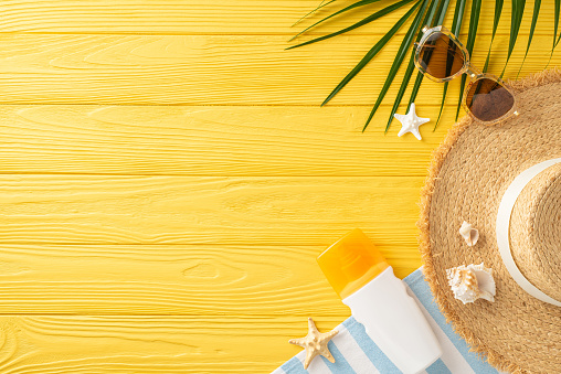 Sun-kissed setup: Overhead view of straw hat, sunglasses, sunscreen bottle, beach towel, palm leaves, shells, and starfish on yellow wooden backdrop, ideal for summer-themed ads