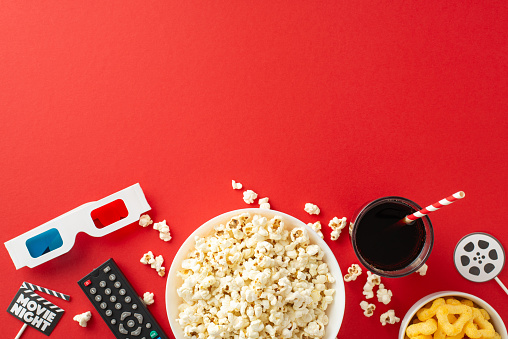 Curate a cozy home theater ambiance with top view of popcorn, snacks, soda, 3D glasses, and remote control. Movie-themed decor on red background allows for text or ads