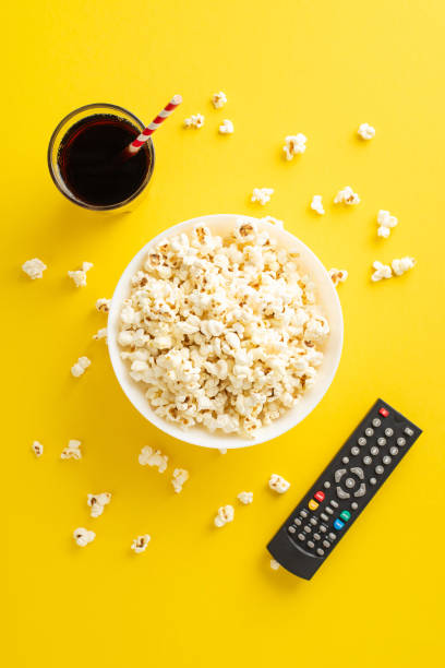 Vertical top view of home cinema gathering featuring savory snack like popcorn, paired with fizzy soda. Set for online streaming with a TV remote. Cheerful movie-themed accessories on a yellow surface vista superior vertical de una reunión de cine en casa con bocadillos salados como palomitas de maíz, combinados con refrescos gaseosos. configurado para transmisión en línea con un control remoto de tv. alegres accesorios con temática de película so