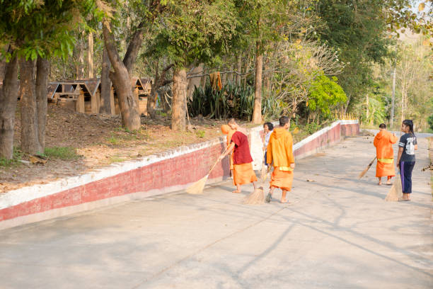 View down and along street with young thai people. There are young temporarily monks who are cleaning street  near and outside of temple Wat Phra Phutthabat Huai Tom in Li District, Lamphun. Street is leading uphill. A young woman and a school girl in school uniform are helping cleaning street with brooms.  In background are many trees