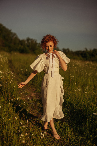 This evocative image depicts a young woman with curly red hair walking through a sunlit meadow during late afternoon. Her vintage-style white dress, with puffed sleeves and a tie at the neck, adds a timeless elegance to the scene. She appears absorbed in the moment, gently touching the wildflowers as she walks by. The warm, natural lighting and the open field background enhance the pastoral and peaceful ambiance, evoking a sense of freedom and connection to nature.