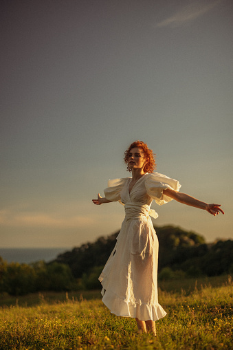 This evocative image depicts a young woman with curly red hair walking through a sunlit meadow during late afternoon. Her vintage-style white dress, with puffed sleeves and a tie at the neck, adds a timeless elegance to the scene. She appears absorbed in the moment, gently touching the wildflowers as she walks by. The warm, natural lighting and the open field background enhance the pastoral and peaceful ambiance, evoking a sense of freedom and connection to nature.