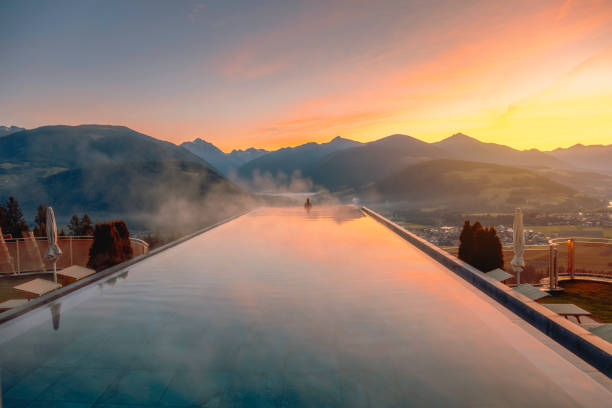Young woman relaxing in the infinity pool with a nature landscape view in Dolomites, Italy As the sun rises over the breathtaking landscape of the Dolomites in Italia, a woman in a bikini finds pure bliss and relaxation in the well-heated infinity pool atop a luxurious hotel. With a radiant smile, she immerses herself in the warm waters, feeling the gentle steam rising around her. The pool's edge seems to blend seamlessly with the horizon, offering an awe-inspiring panorama of the surrounding alpine scenery. Surrounded by the majestic peaks of the Dolomites, she feels a profound sense of peace and tranquility wash over her. In this moment, high above the world below, she is truly in harmony with nature, cherishing every precious moment of serenity in this alpine paradise. vista posterior stock pictures, royalty-free photos & images