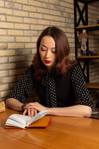 In a cozy cafe, a stylish young woman in a black polka dot blouse, long brown hair, and red lipstick, enjoys a serene reading session, immersed in a captivating book.