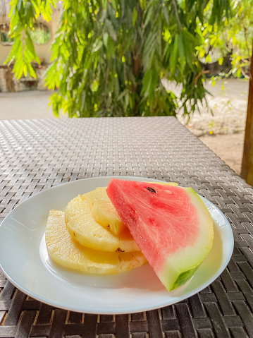 A close-up view of a white plate with juicy pineapple and watermelon slices, set on an outdoor table with a tropical backdrop.