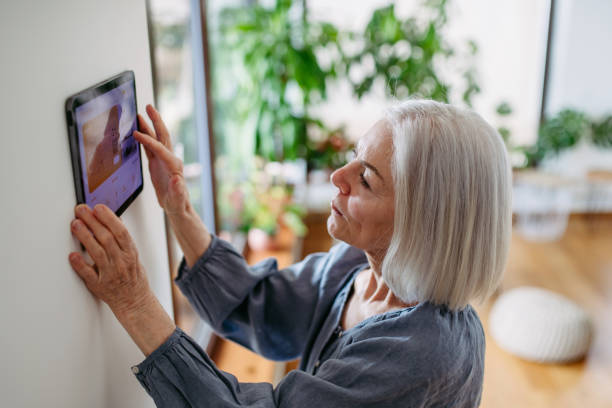 Mature woman adjusting smart thermostat, touch screen of smart home device. Older woman using smart technology at home. Mature woman adjusting smart thermostat, touch screen of smart home device. Older woman using smart technology at her home. home automation stock pictures, royalty-free photos & images