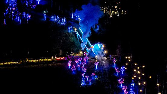Overhead Night View Of A Colorful Light-Festooned Train Emitting Steam As It Travels Through A Dark, Tree-Lined Area.