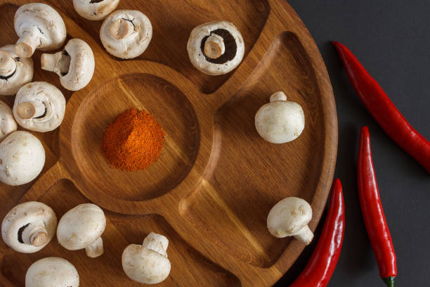 Still life of vegetables. A wooden plate on which contains champignons, pods of acute red pepper and a pile of milled pepper. Dark background. View from the top. Culinary concept.