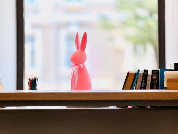 A pink easter bunny decoration sitting on a table with books and window on background