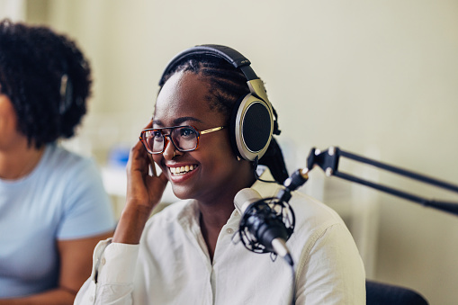 A focused African-American female podcaster with a warm smile, wearing headphones and glasses, radiates confidence while conducting a podcast episode in a modern studio setting.